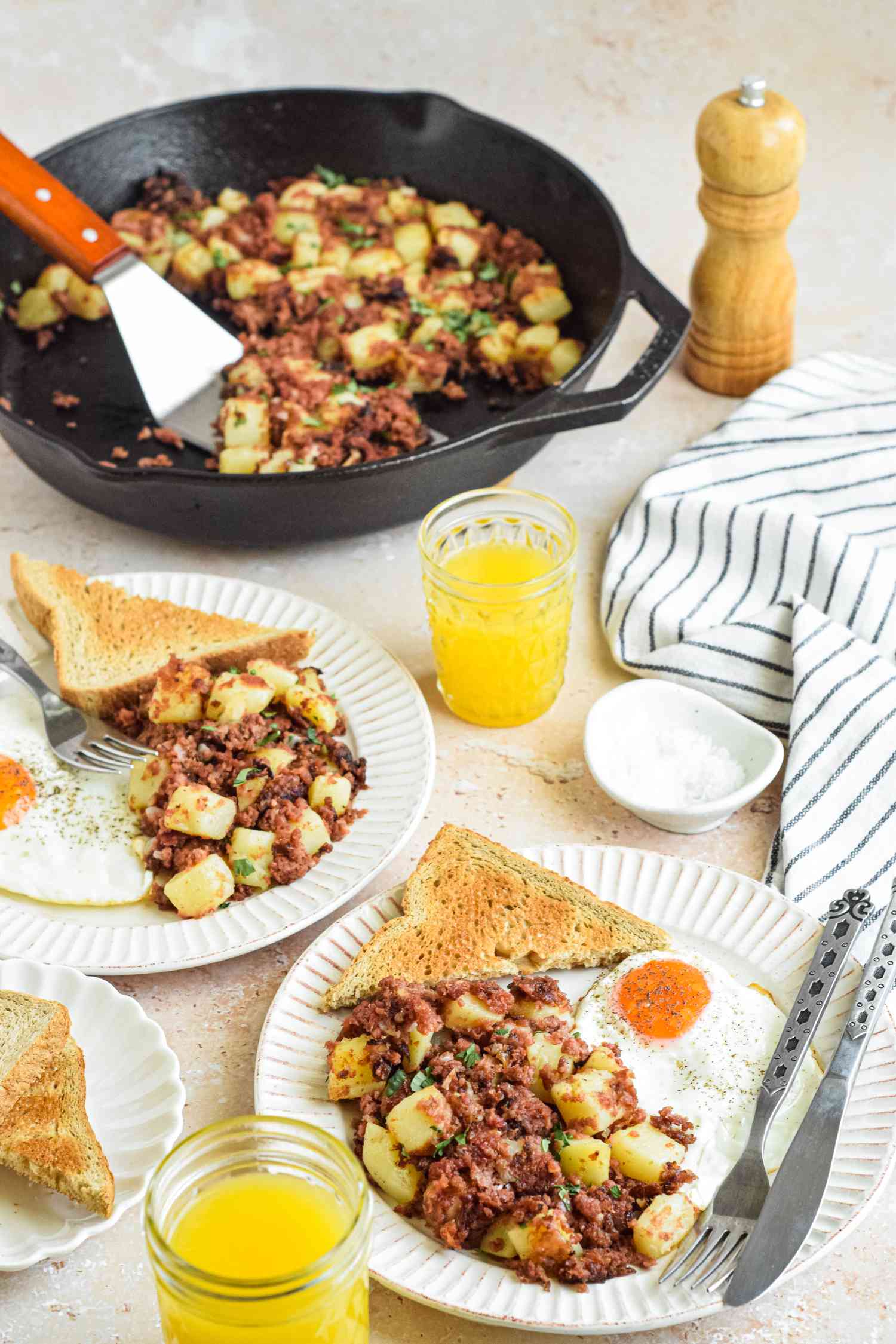 Corned Beef Hash Served on Two Plates with Toast and Sunny Side Eggs, Surrounded by Glasses of Juice, a Plate of Toast, and More Corned Beef Hash in a Cast Iron Skillet