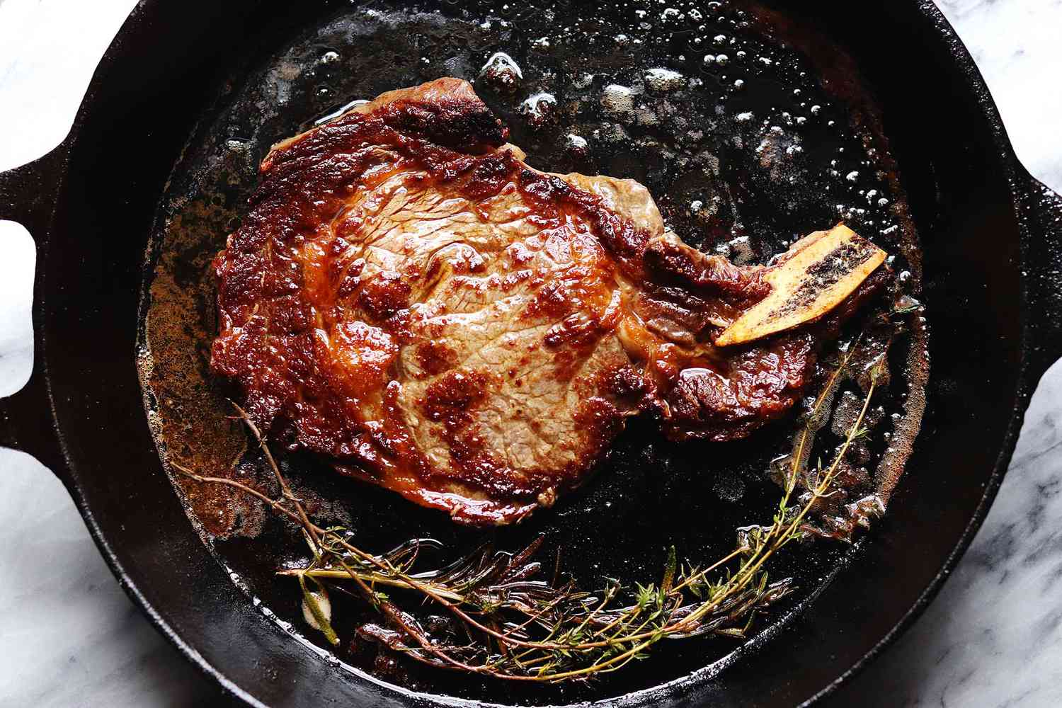 A cast iron skillet with steak cooked on the stove. Oregano sprigs are at the bottom of the pan. Oil and juices are visible in the pan.