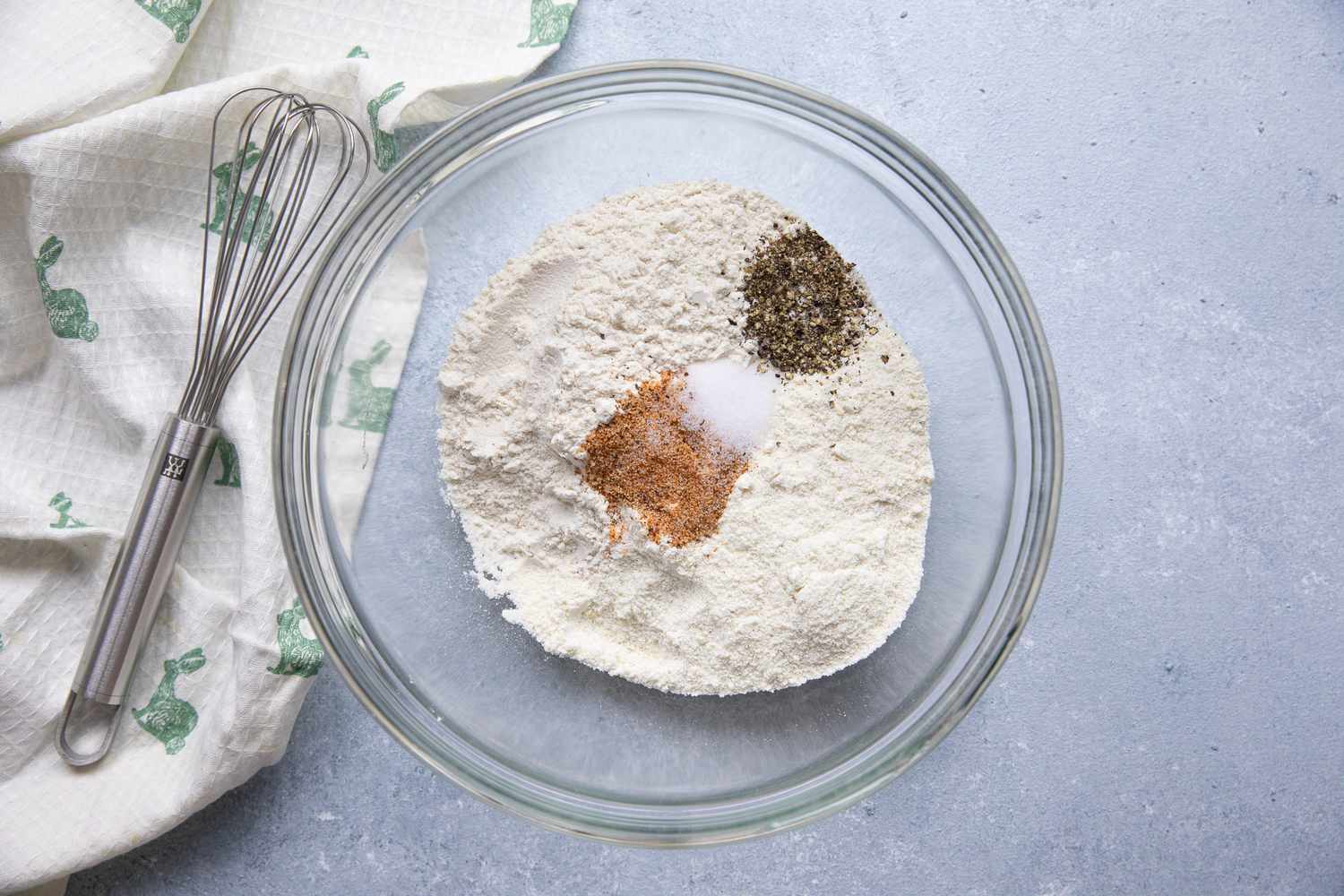 Bowl of Dry Dredging Ingredients (Flour, Corn Meal, and Spices) Next to a Kitchen Towel with a Whisk for NOLA Oyster Po' Boy Recipe