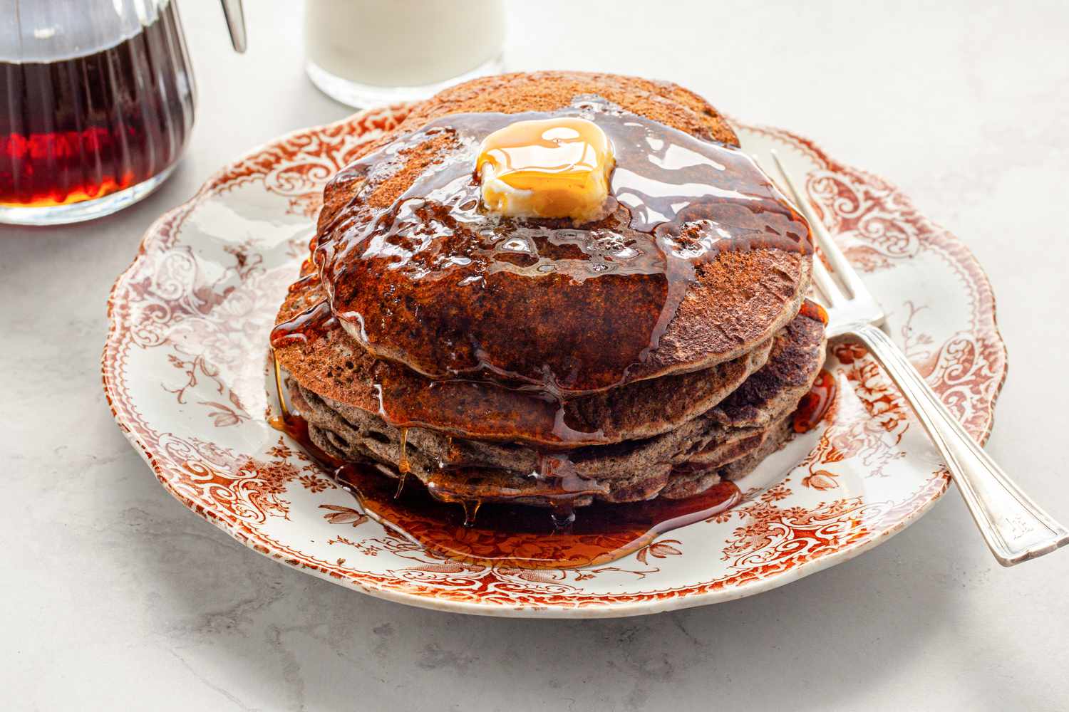 Stack of Buckwheat Pancakes Topped with Maple Syrup and Some Butter, and Next to It, a Glass of Milk and a Container of More Syrup