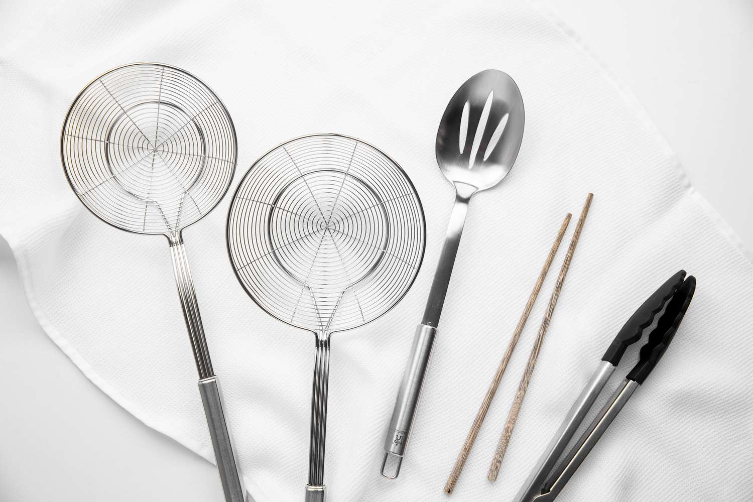 Tools for Deep Frying (L to R): Two Spiders, Slotted Spoon, Chopsticks, and Tongs, All on a White Kitchen Linen 