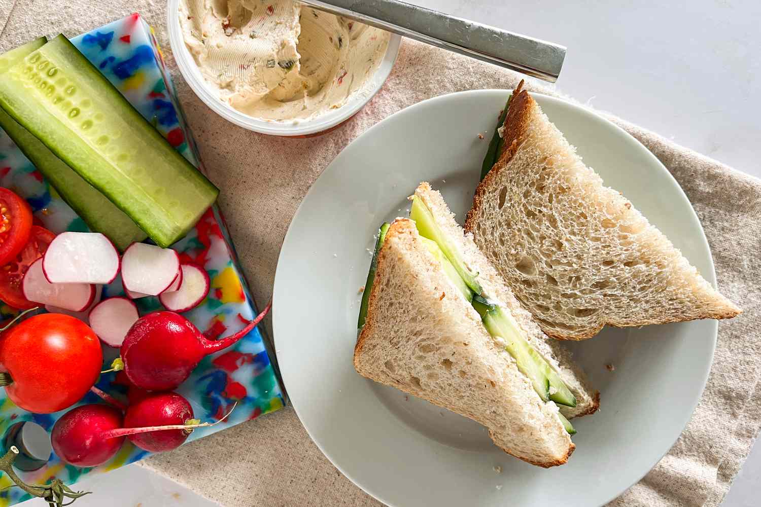 halved 1-minute cucumber sandwiches on a plate next to a cutting board with sliced vegetables and a opened tub of Tilamook Cream Cheese