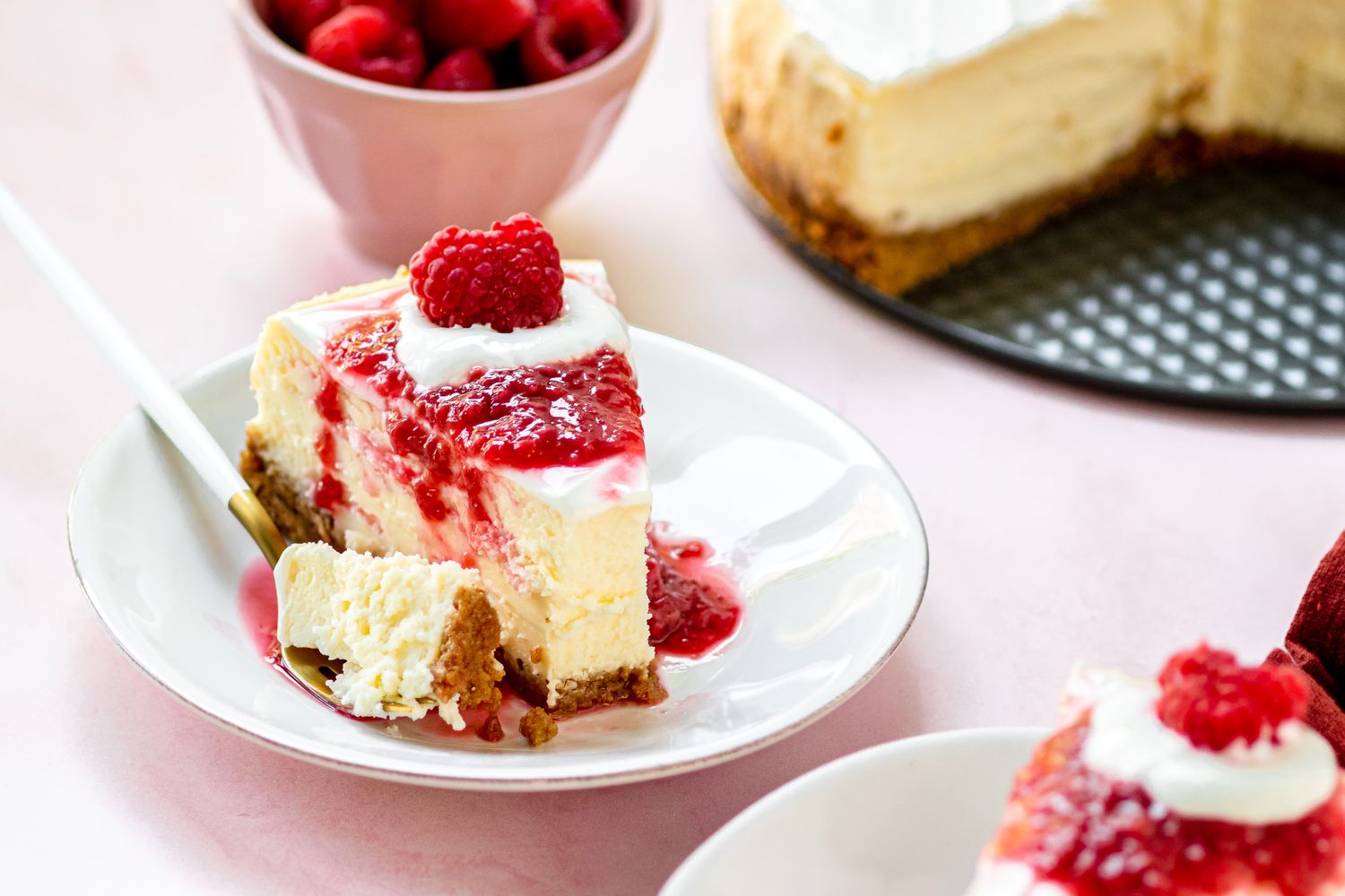A plate with a slice of New York cheesecake topped with raspberry sauce and a forkful of cheesecake set on the plate.
