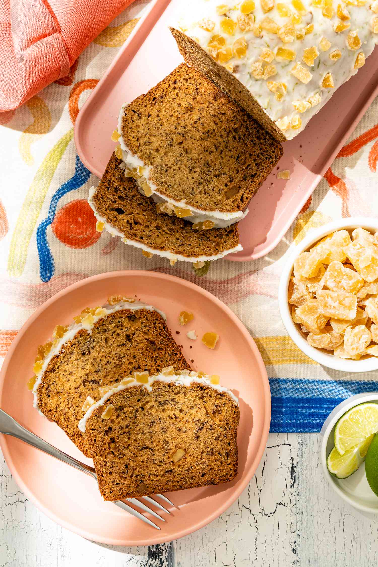 Dark and Stormy Banana Bread on a Platter With Part of the Loaf Cut Into Slices, and in the Surroundings, More Slices of the Loaf on a Plate With a Fork, a Bowl of Candied Ginger, Bowl of Lime Slices, and a Pink Table Napkin, All on a Decorative Table Cloth