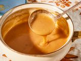 Angled view of a white pot of gravy sitting on a wooden trivet with a ladle with some gravy and all on a white and brown embroidered table cloth