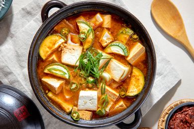 Overhead View of Doenjang Jjigae in a Clay Pot on a White Kitchen Towel and Surrounded by a Bowl of Rice, a Small Bowl of Sauce, and a Wooden Spoon