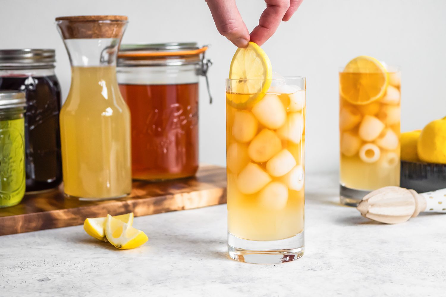 Lemon Slice Added to Glass of Arnold Palmer With Ice. In the Background, Another Glass With Tea, Ice, and Lemonade, a Bowl of Lemons, and a Citrus Reamer, and on the Left, Two Jars (One With Lemon Juice and Another With Black Tea) on a Wooden Board and Two Lemon Wedges.