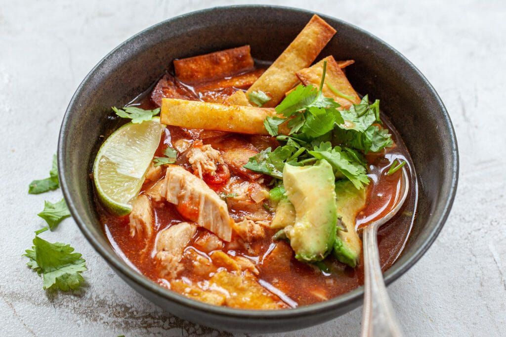 chicken tortilla soup served in a bowl with a spoon, topped with tortilla chips, lime, and cilantro