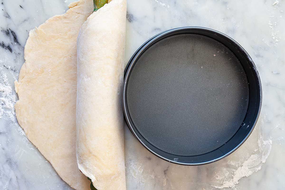 Mile high quiche crust dough is on a marble countertop and is rolled around a rolling pin. An empty springform pan is to the right.