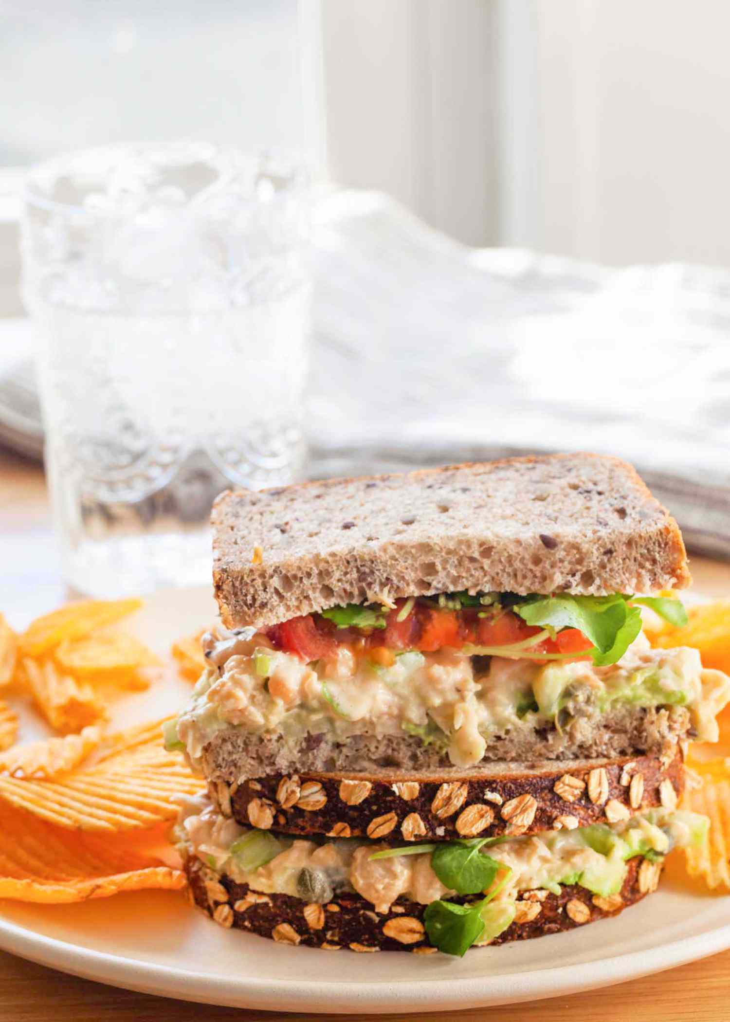Side view of two easy chickpea sandwiches stacked on a plate with chips. The filling includes chickpea salad, sprouts and tomato.