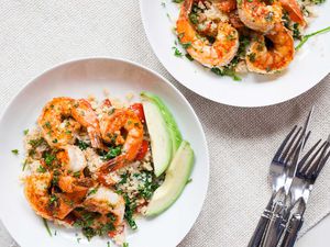 Two white plates on a white table with a stack of forks and knives to the right. Each plate has large shrimp, avocado slices, herbs and cauliflower rice inside.