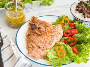 Side view of lenten fish with garlic sauce on a plate with a blue edge. A blue striped napkin and a black fork are to the left of the plate. The large fillet is crispy and pink on the outside. Lettuce and cut red and orange peppers are to the right of the snapper. A bowl of rice and black beans is to the upper right of the image. A jar with a spoon inside has chillo al Ajuillo inside. Behind is a platte with more lettuce and snapper.