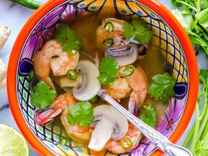 Thai soup with shrimp in a brightly patterned bowl with a silver spoon inside. Sliced lime, ginger, peppers and cilantro stems suround the bowl of light broth, shrimp, mushrooms and cilantro.