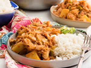 Horizontal view of rimmed grey plate with heaping portion of Puerto Rican Cod Fish. White rice and sliced avocado are on the plate along with a fork. Another plate is to the upper right and a dutch oven is partially in view in the upper center. A blue casserole dish is heaped with white rice and is above the plate to the left. A pink and blue patterened linen is under the plate and rice dish.