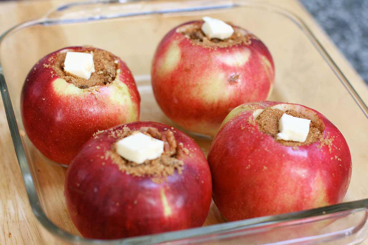 Four hollowed out apple stuffed with a sugar stuffing mixture and topped with a cut of butter, resting in a baking dish