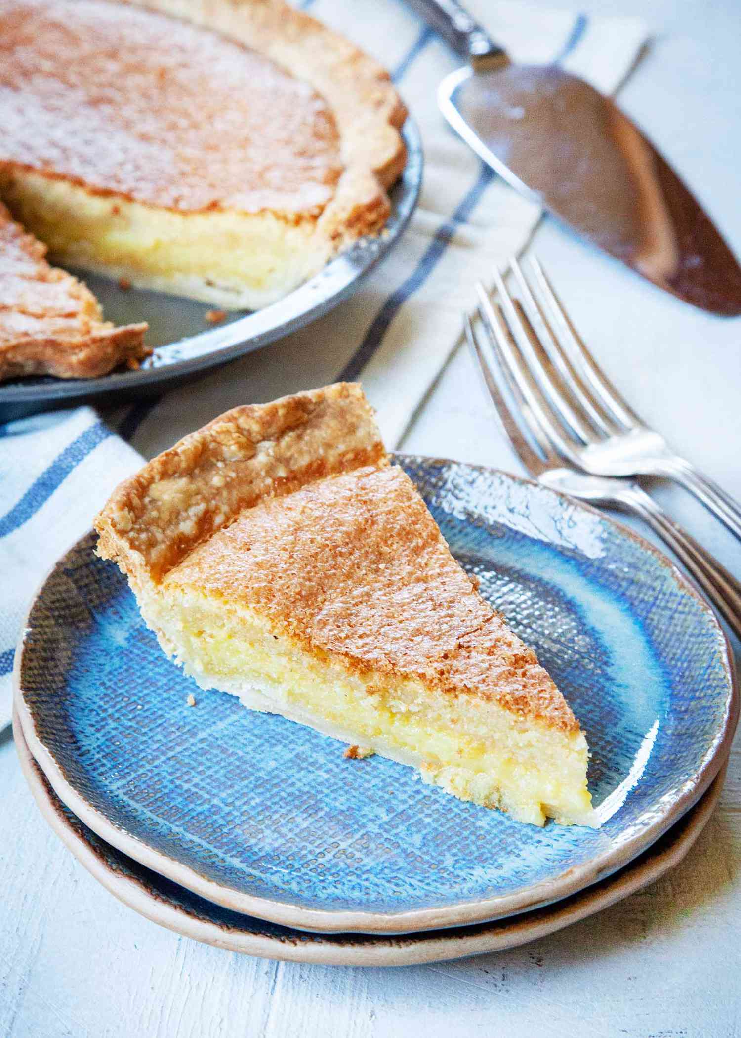 A slice of chess pie on stacked blue plates. Forks are set to the right and the rest of the pie is behind it.