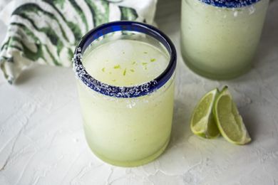 Frozen margarita in a blue rimmed glass with limes in front and another glass and linen behind it.
