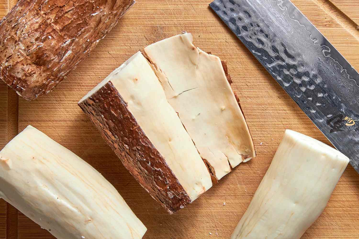 Yuca / Cassava Root With the Outer Skin Slightly Removed Next to More Yuca Roots (Some With Skin Removed and Some With Skin Intact) and a Knife, All on a Cutting Board