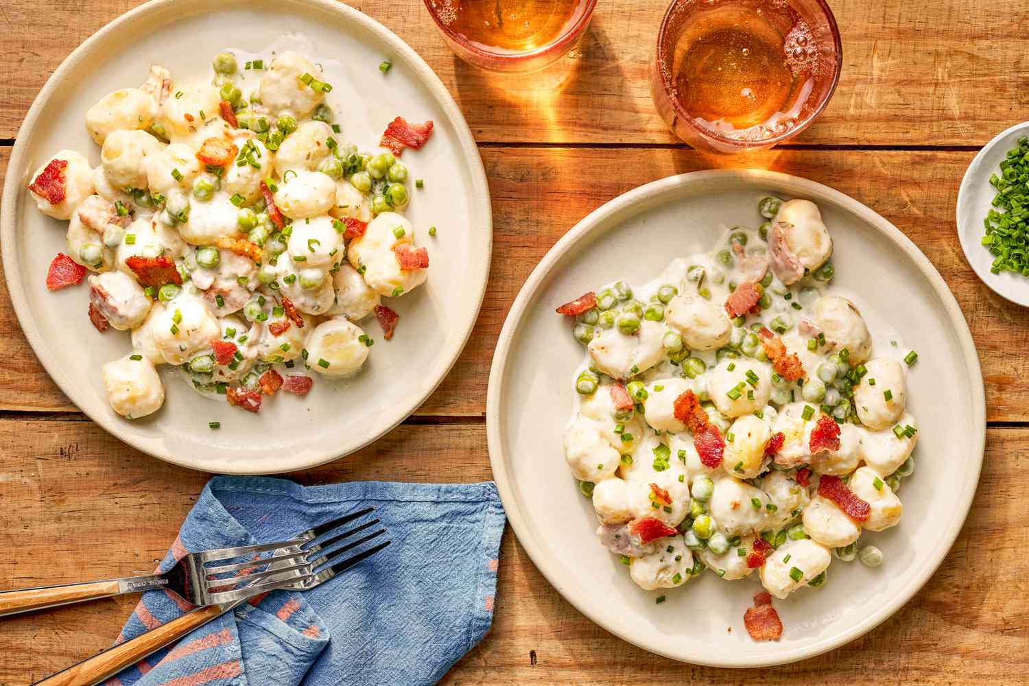 Creamy Skillet Gnocchi With Bacon and Peas served in two plates at a table setting with two drinks, a small plate of chopped chives, and forks on a blue and pink table napkin