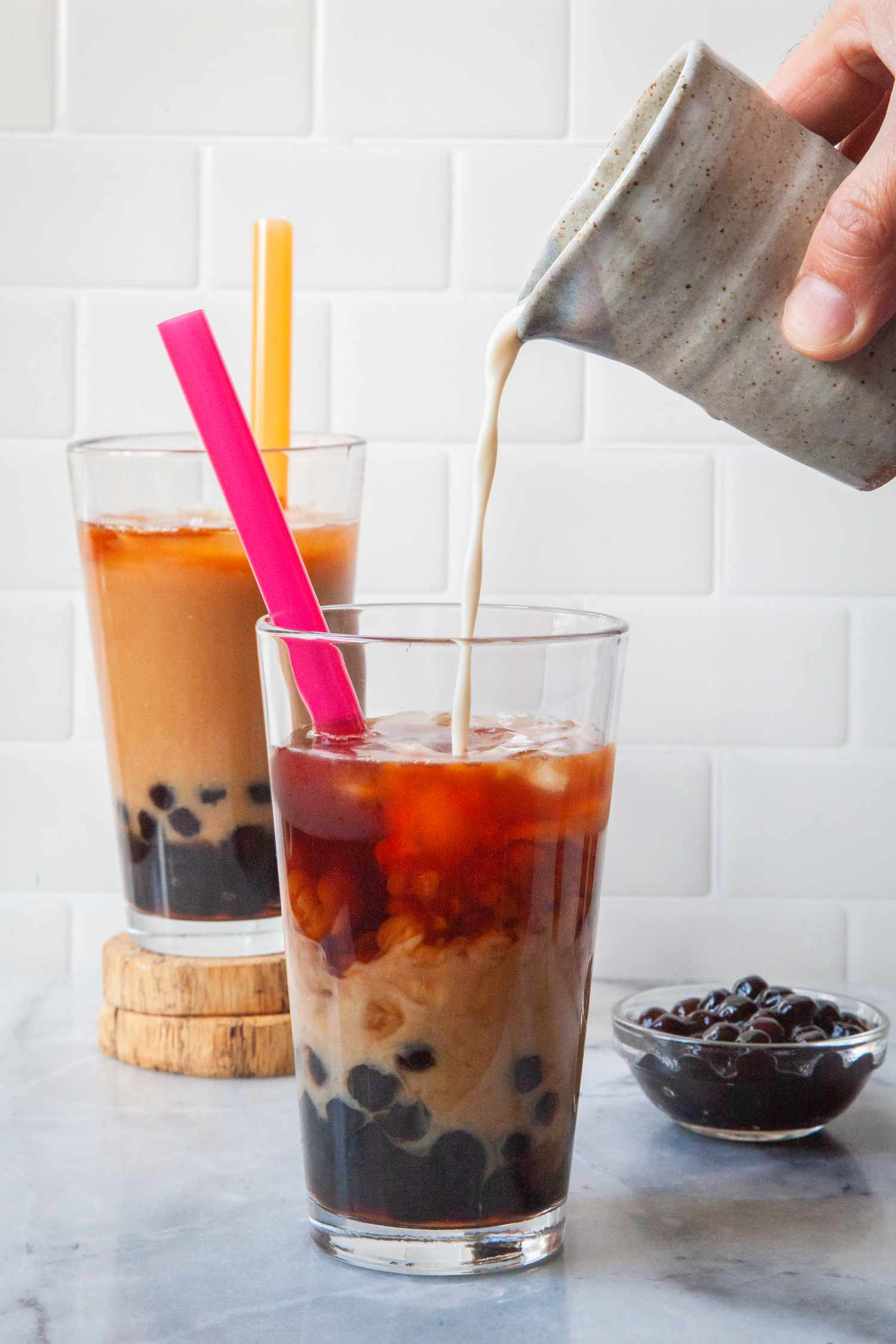 Milk From a Cup Poured Into a Glass With Tapioca Pearls and Black Tea, and in the Surroundings, a Glass of Bubble Tea in the Background and a Small Bowl of Boba