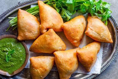 Vegetarian Samosas on a bed of herbs with one samosa filled with potato and peas alongside a cilantro mint chutney or dipping sauce in a small wooden bowl..