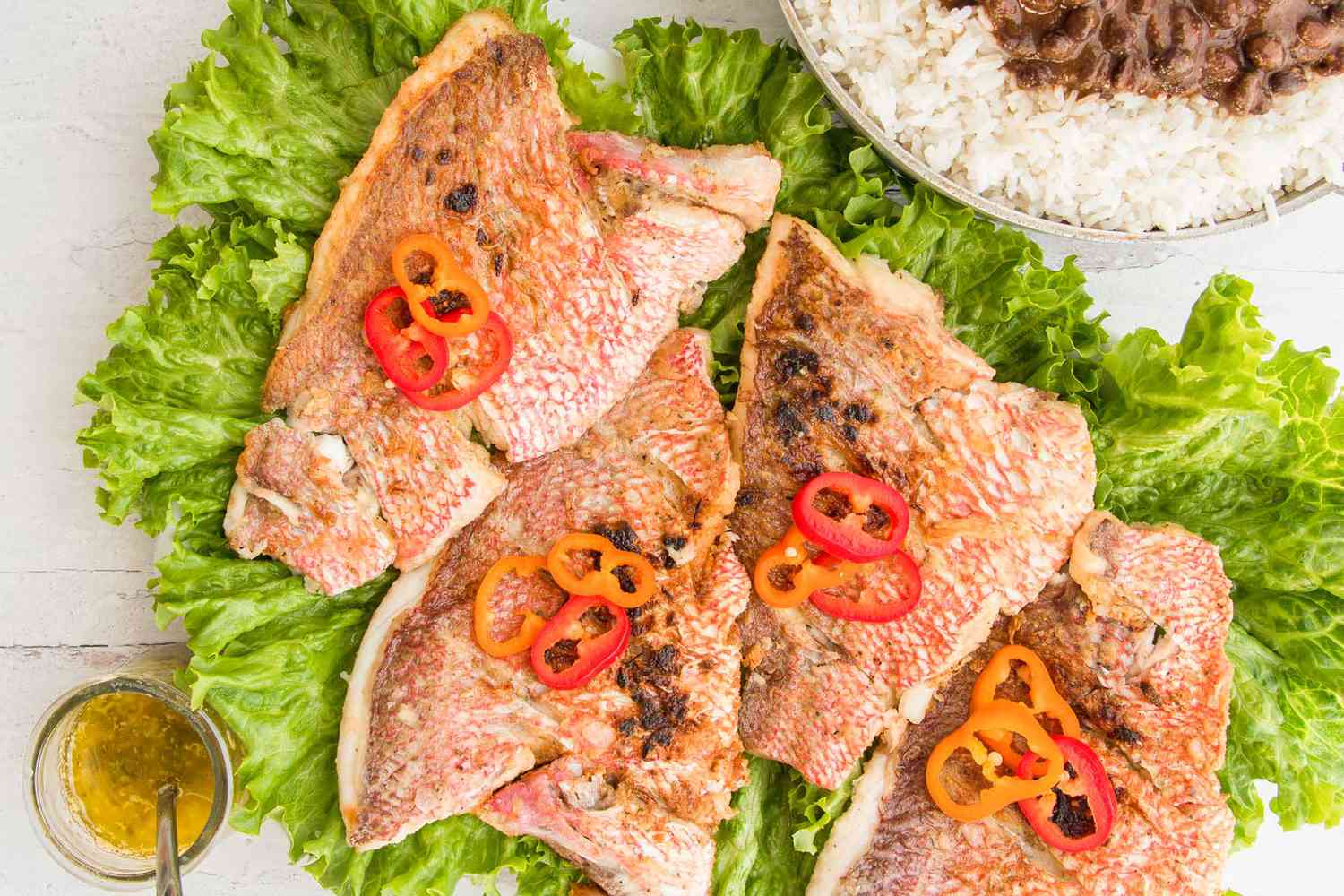 Overhead view of a platter of homemade red snapper fillets layered on lettuce. A small jar of garlic sauce has a spoon inside. A large bowl of rice and black beans is in the upper right corner.