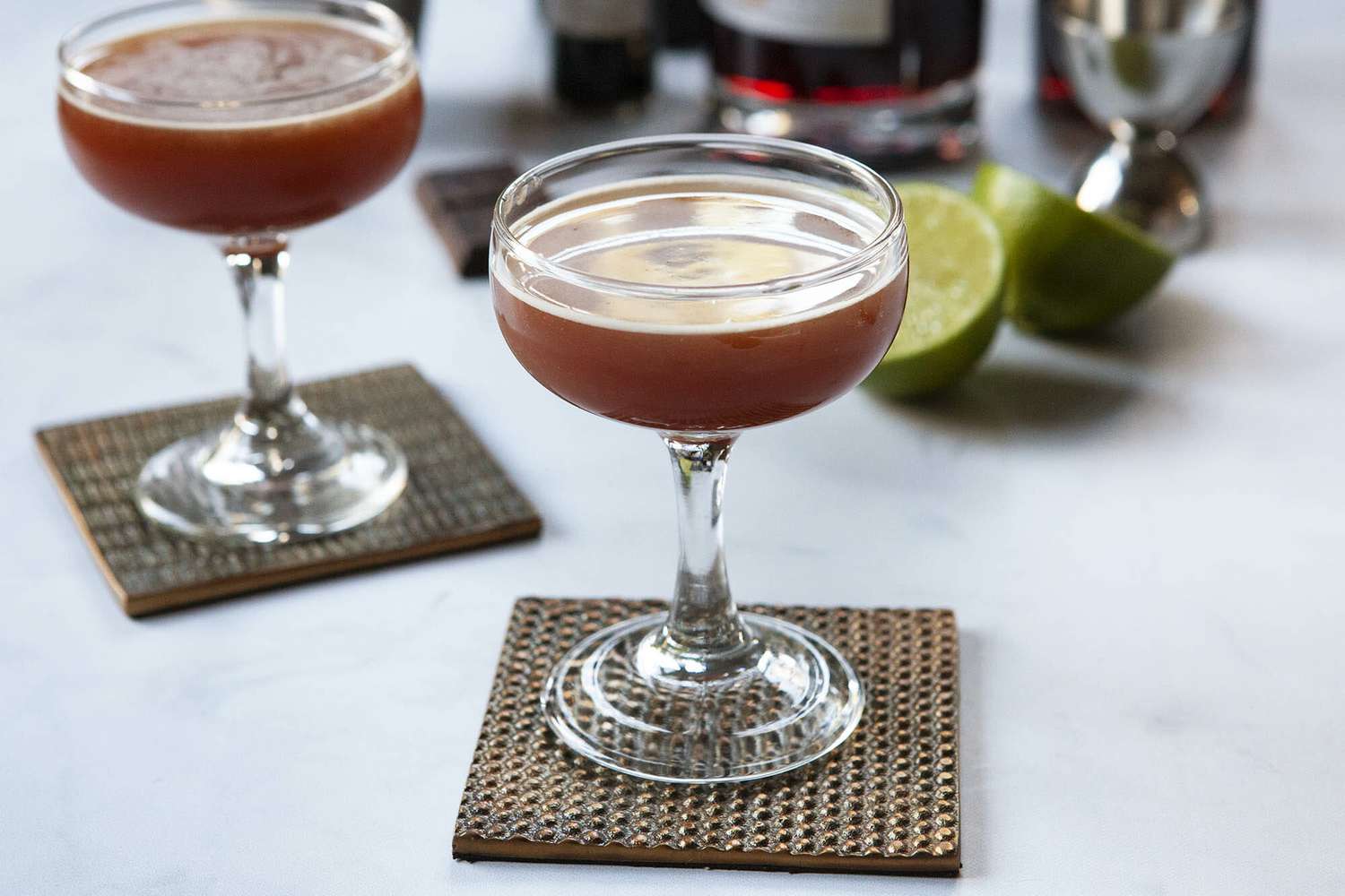 Sidecar drink with chocolate in a coupe glass with a second glass behind it. Ingredients for the cocktails are behind the glass.