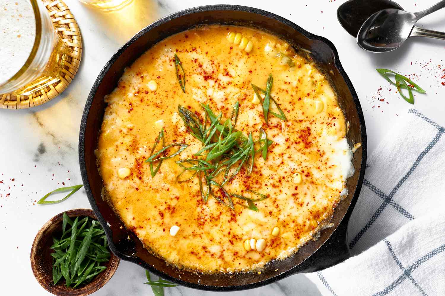 Overhead View of Korean Corn Cheese Topped With Sliced Green Onions and Gochugaru, and in the Surroundings, a Bowl of Thinly Sliced Scallions, a Glass of Water, and a White Kitchen Towel