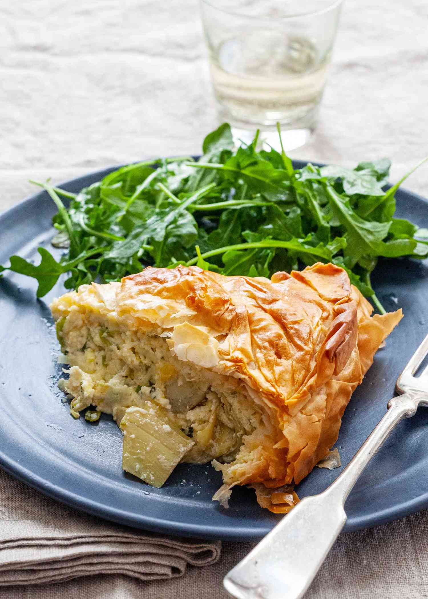 Side view of a slice of spring pie on a blue plate with a golden, flaky crust and topping. Chopped artichokes are visible in the cheesy filling. A bright arugula salad is behind the pie on the plate. A glass of white wine is behind the plate. Linens are under the plate and a fork is set to the right of the pie on the plate.