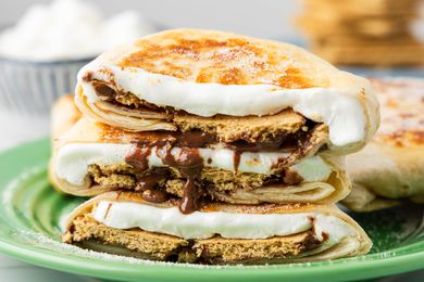 Cross-Section Cut of a S'mores Crunchwrap on a Plate, and in the Background, a Bowl of Mini Marshmallows and a Plate With a Stack of Graham Crackers