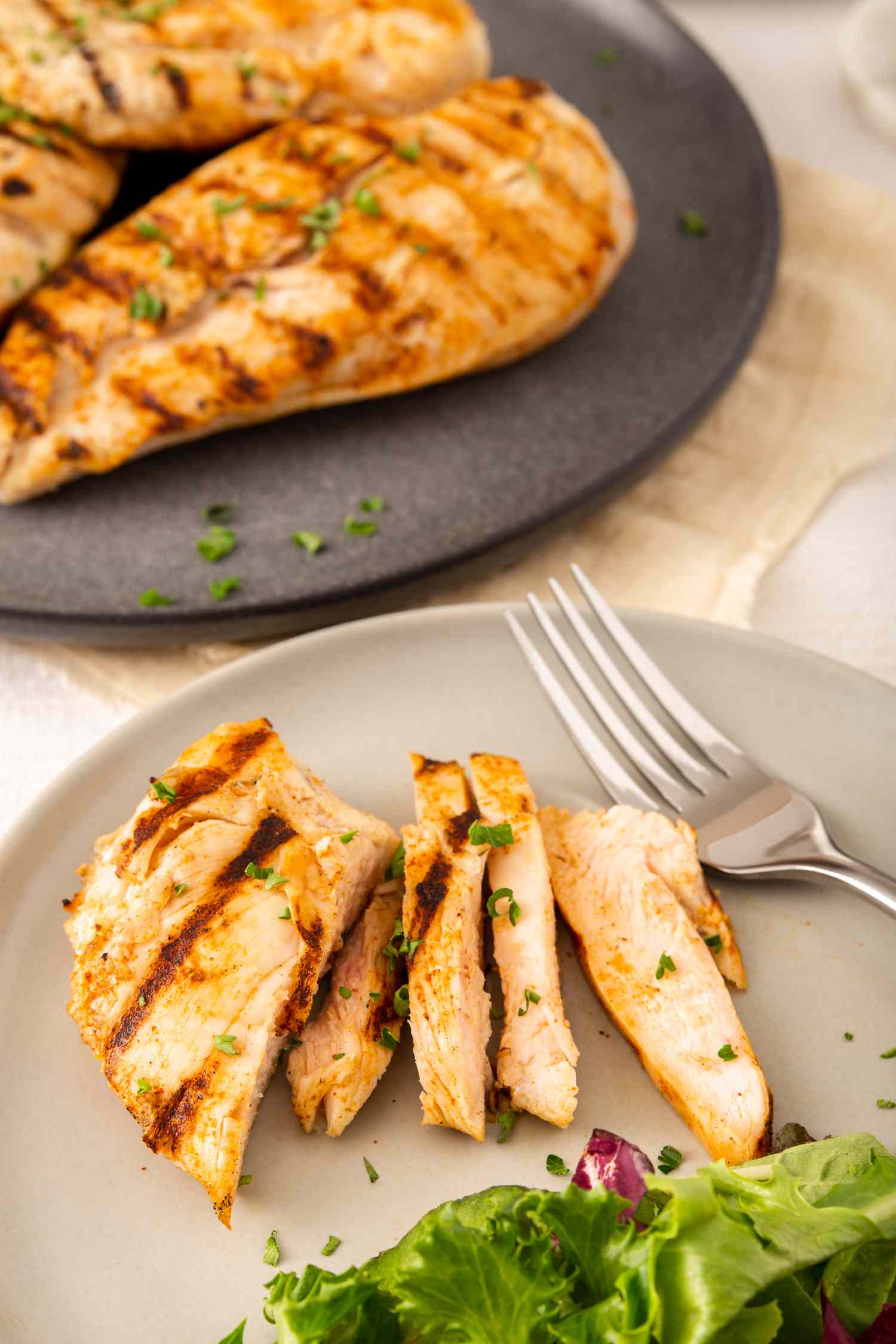 Grilled chicken breast sliced on a plate and two whole breasts behind it on another plate.