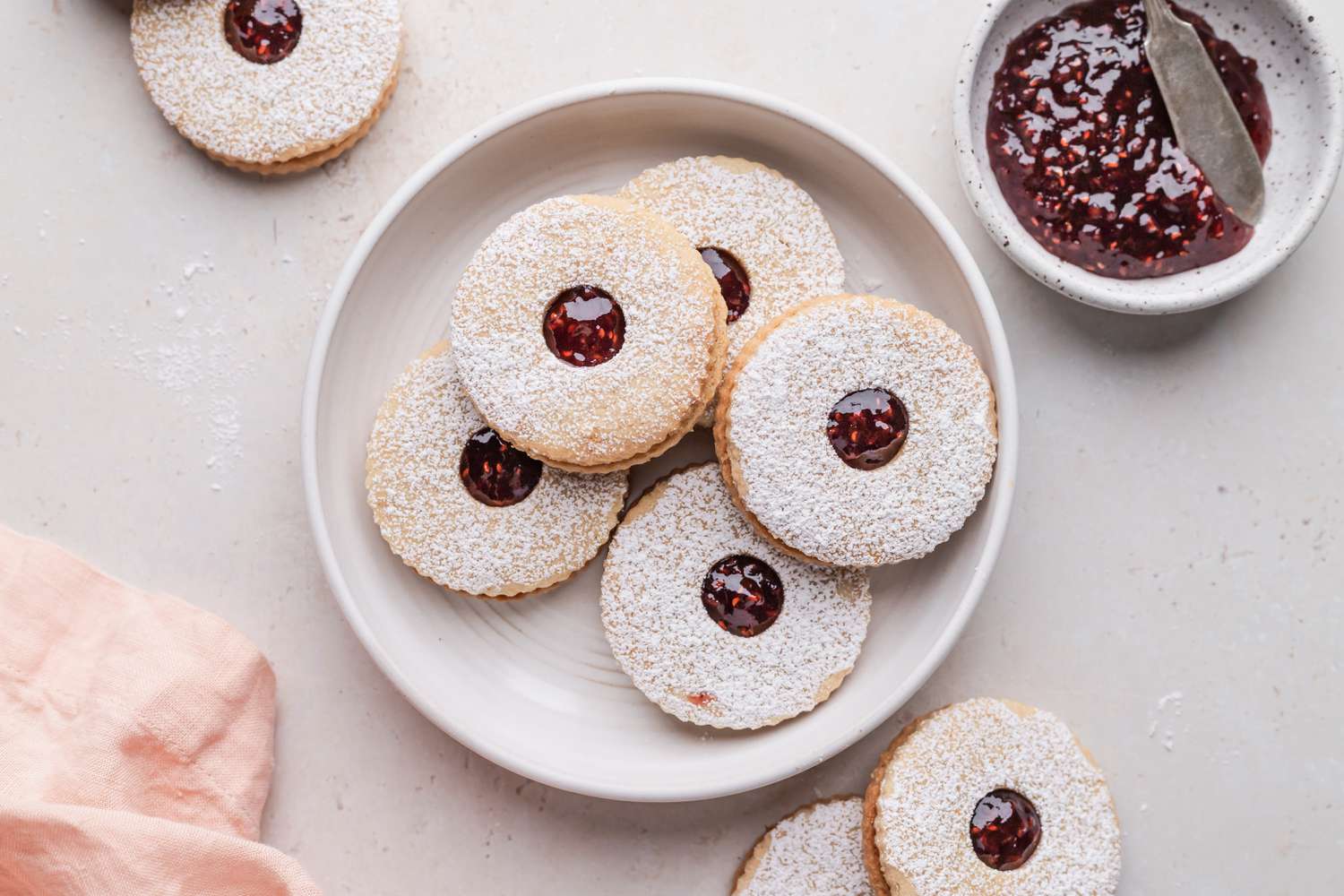 Jam filled sandwich cookies on a plate with additional cookies and jam set around the plate.