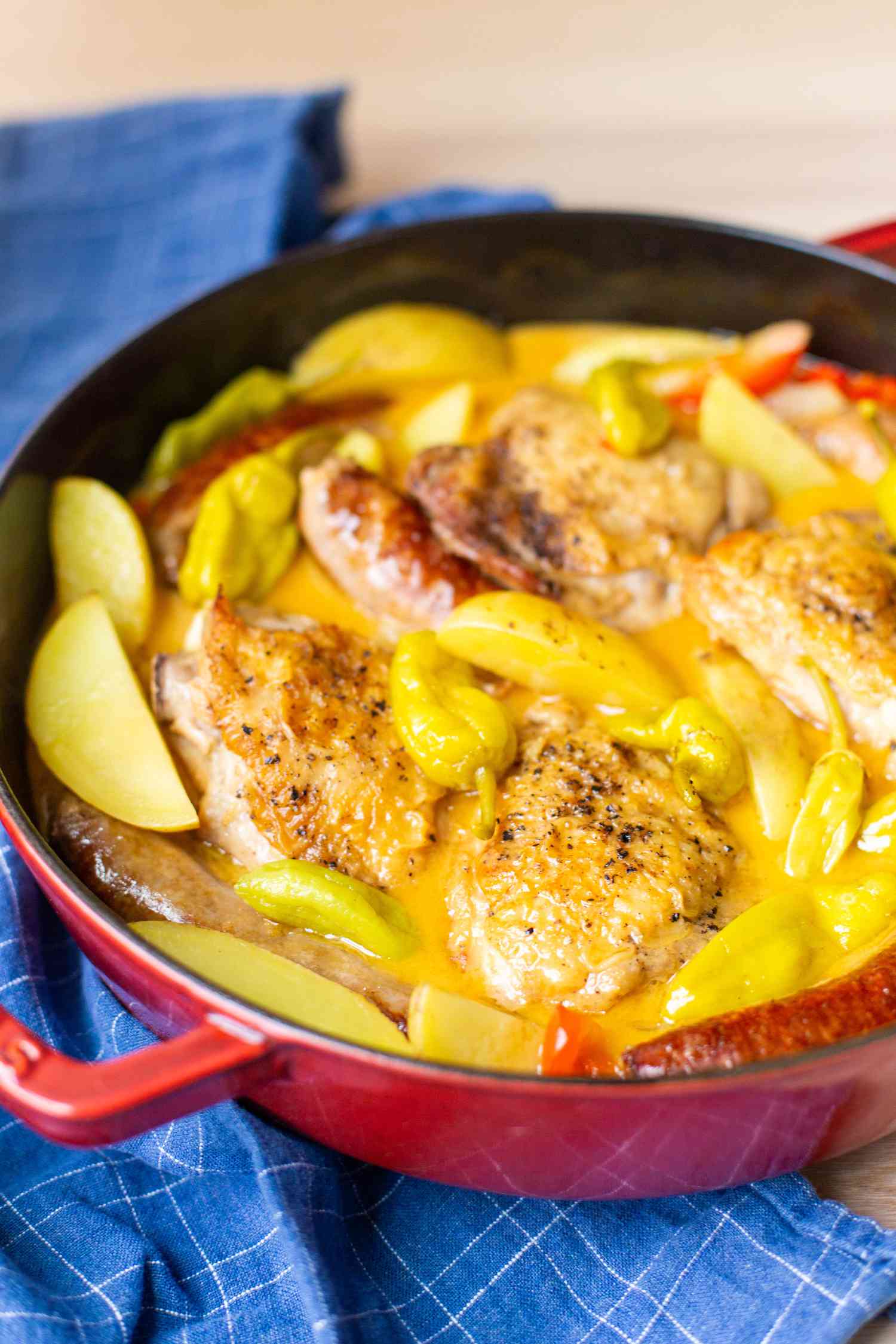 Close-up: Pot of chicken scarpariello in a Dutch oven next to a blue kitchen towel, both on a wooden countertop