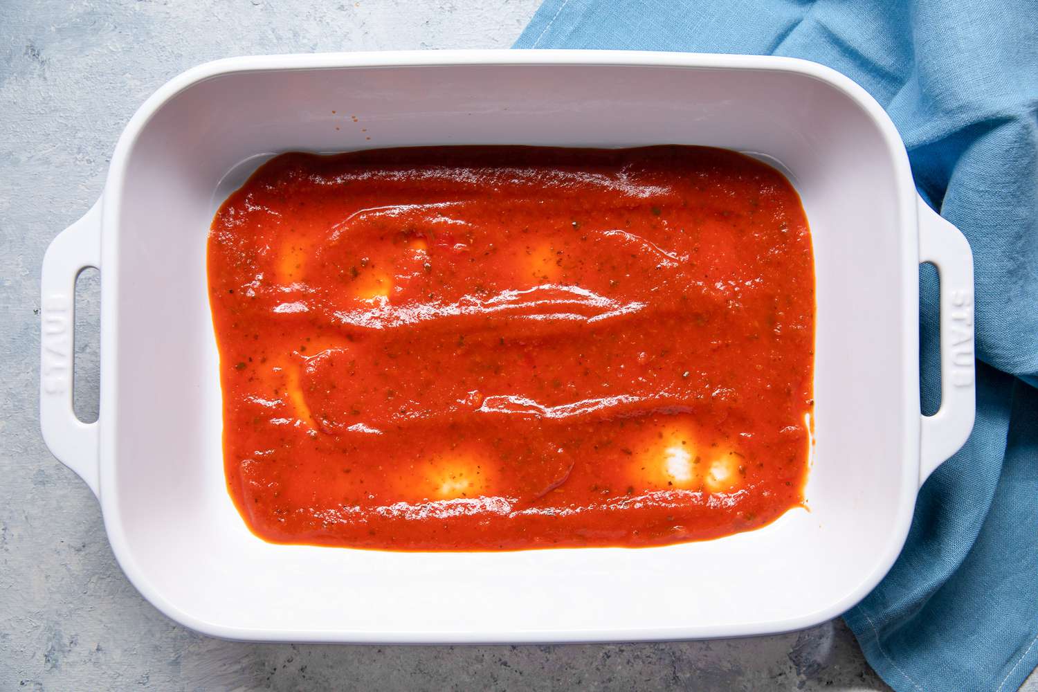 Layer of Marinara Sauce Added to Casserole Dish Next to a Sky Blue Kitchen Towel for Pepperoni Pizza Lasagna