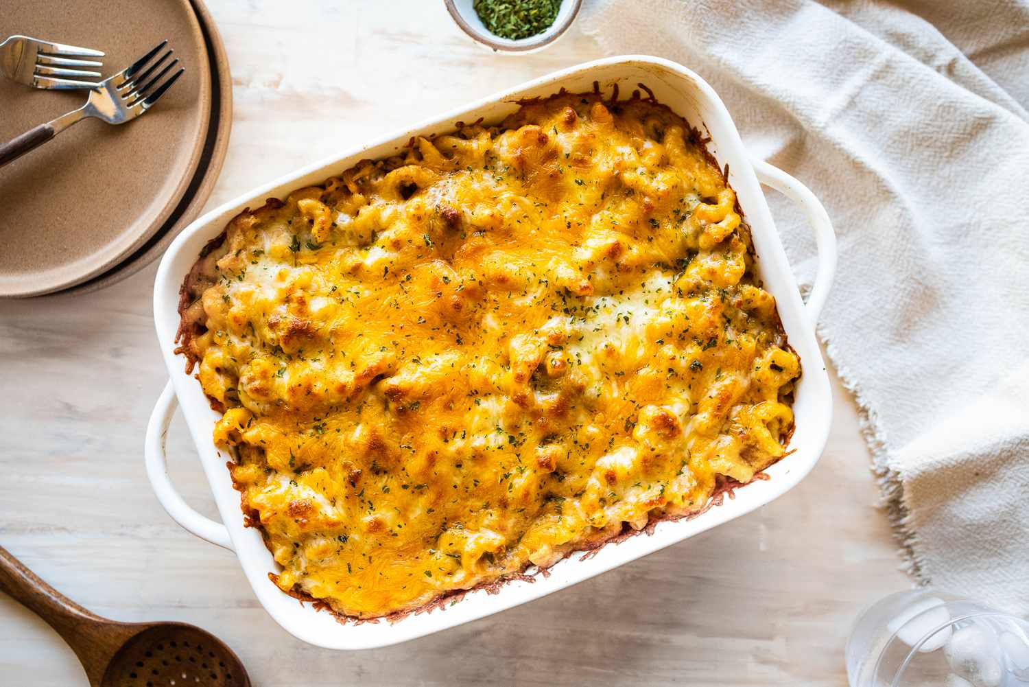 Cheeseburger Casserole in a Casserole Dish at a Table Setting (a Stack of Plates with Two Forks, a Spoon on the Counter, Bowl of Dry Parsley, a Table Napkin, and a Glass of Ice Water)