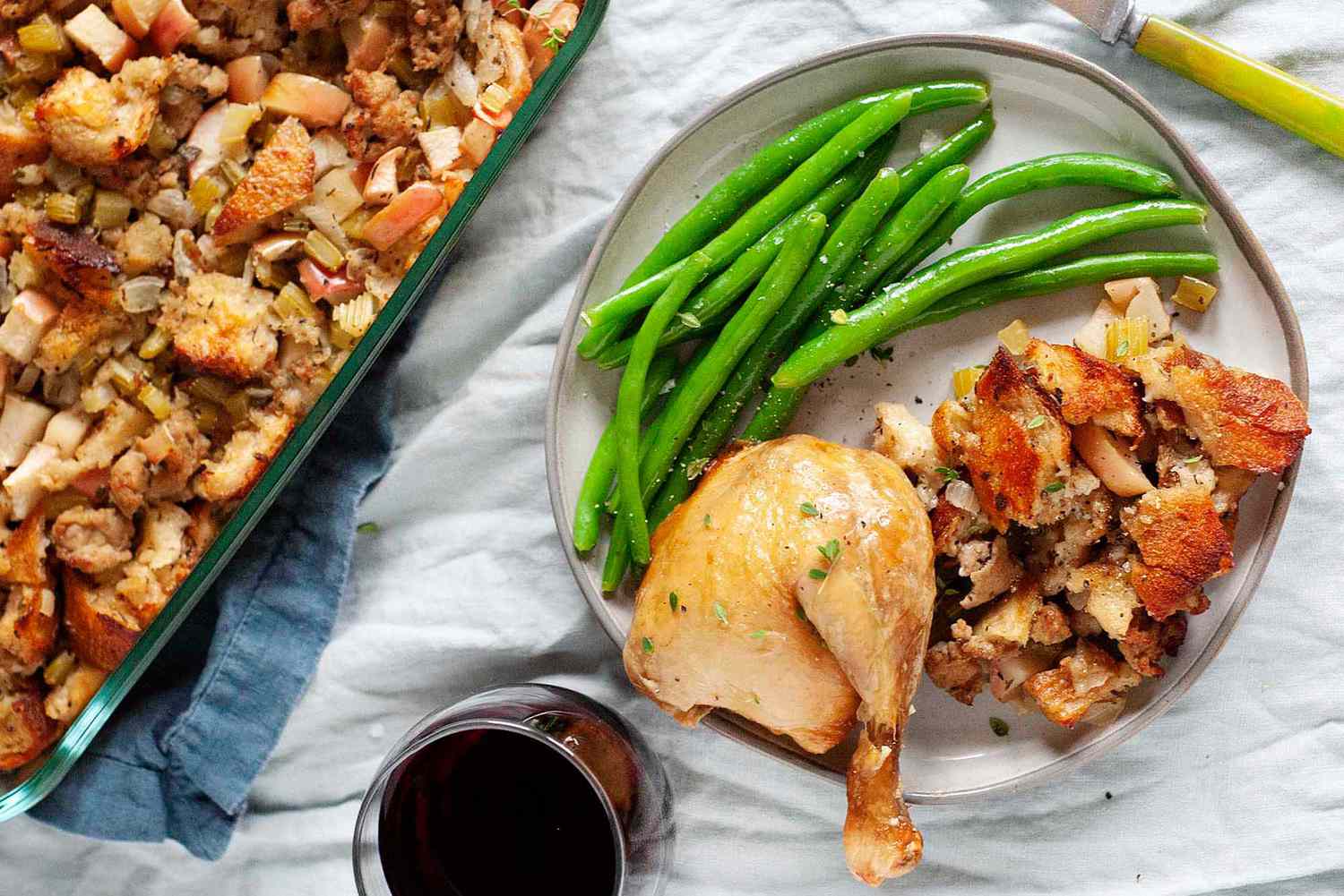 A baking dish with classic sage, sausage and apple stuffing next to plate of stuffing, chicken leg, and green beans. There is a glass of red wine nearby. 