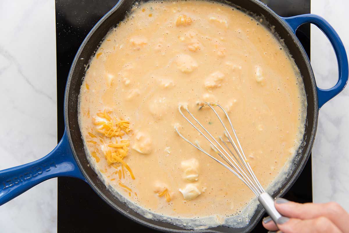 A black cast iron skillet will spices, milk and cheese being whisked together with a wooden spoon for easy homemade queso