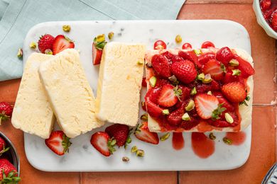 Overhead View: Strawberries and Cream Semifreddo Slices Cascading on a Platter Next the Rest of the Uncut Semifreddo on the Same Platter. Semifreddo Is Topped With Halved Fresh Strawberries and Strawberry Compote. In the Surroundings, a Bowl of Fresh Strawberries, a Stack of Plates, and a Blue Table Napkin.
