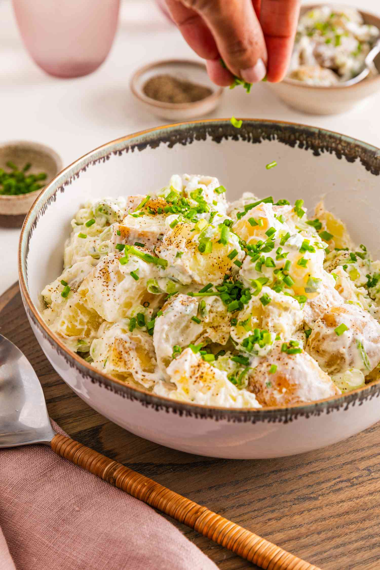 Sliced Chives Sprinkled on a Bowl of Sour Cream and Onion Potato Salad, and in the Surroundings, a Bowl of Seasoning, a Glass of Water, a Bowl of Chives, Stack of Plates With Utensils, and a Blush Table Napkin