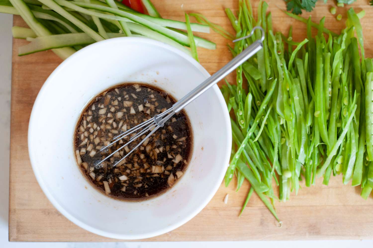 Mixing the dressing in a white bowl with sliced vegetables set next to it for a vegetable rice noodle salad.