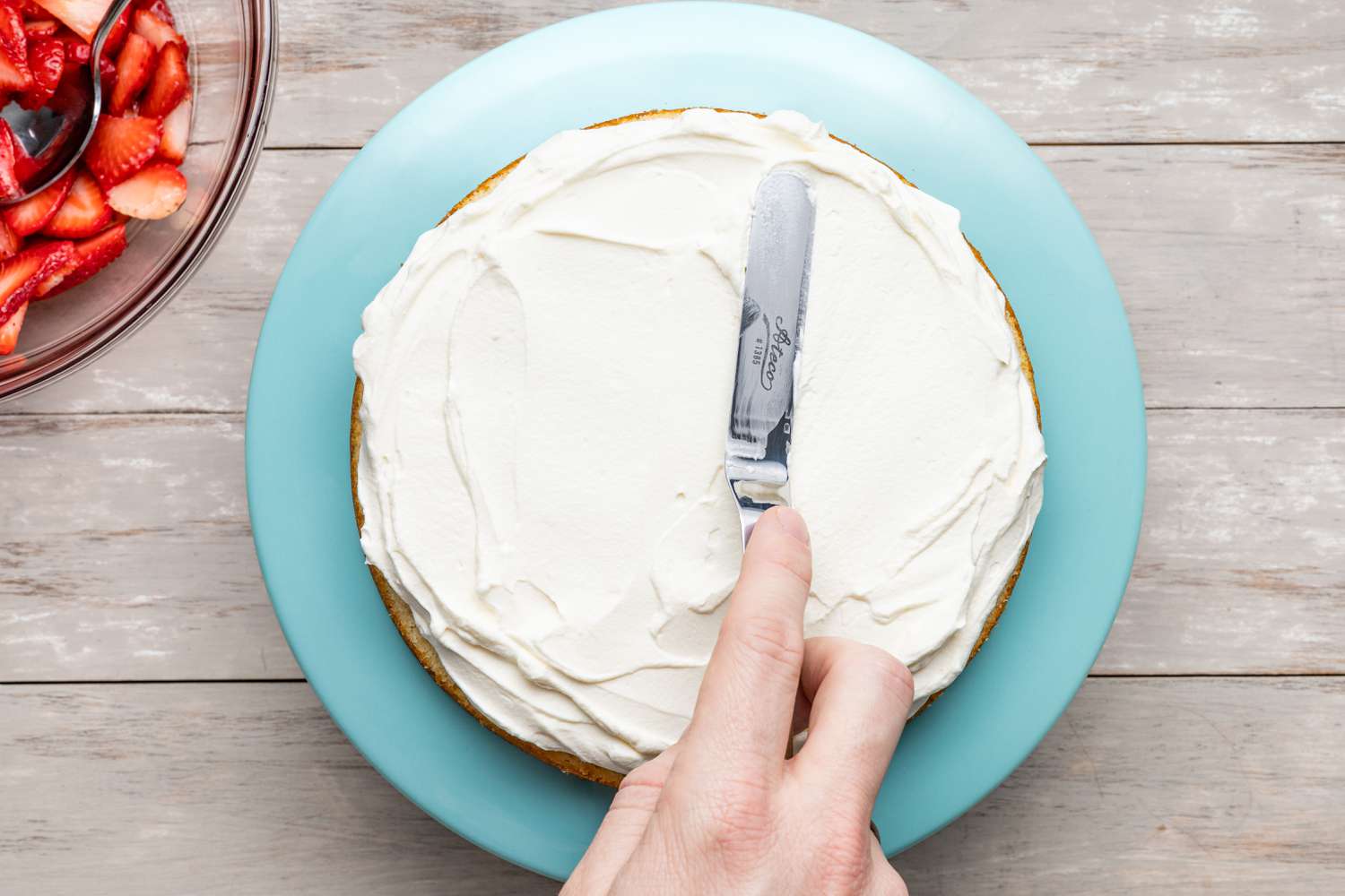 Cream Cheese Whipped Cream Frosting Spread on a Layer of Cake Using an Offset Spatula (Cake on a Cake Stand, Which Is Next the Bowl of Strawberries)