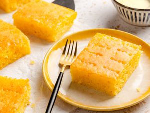 Angled view of a slice of orange poke cake and fork on a white and yellow plate with four slices of cake to the side along with a bowl of glaze
