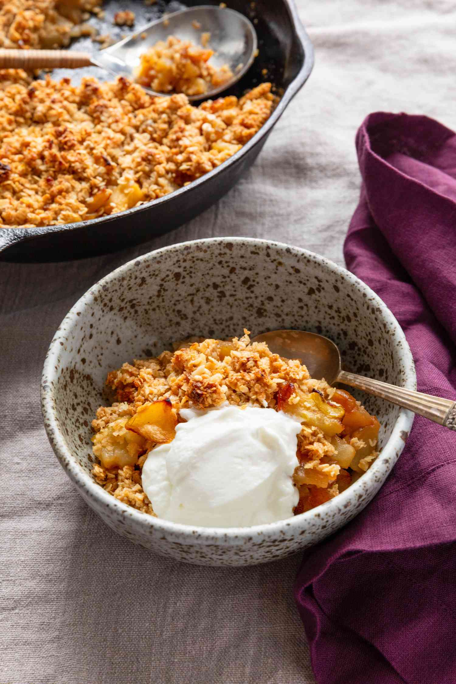 Bowl of 3-ingredient apple crisp with a dollop of yogurt and in the surroudings, a cast iron skillet with more apple crisp and a purple kitchen towel