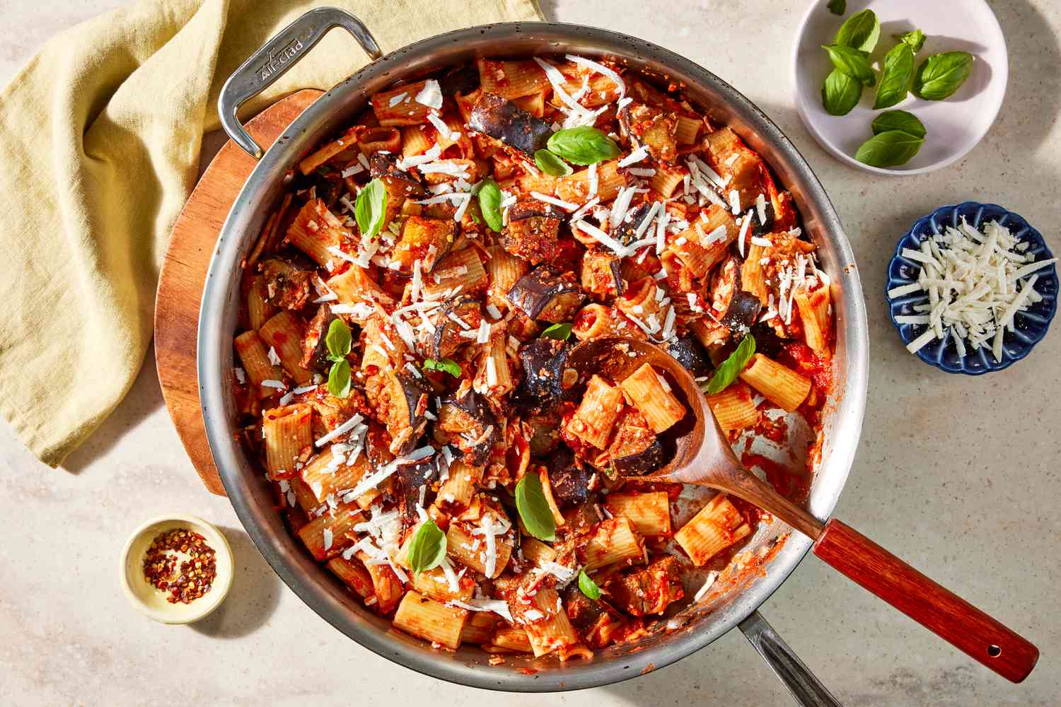 pot of pasta alla norma with a serving spoon and surrounded by bowls of toppings (one with basil leaves, another with shredded cheese, and last with crushed peppers)