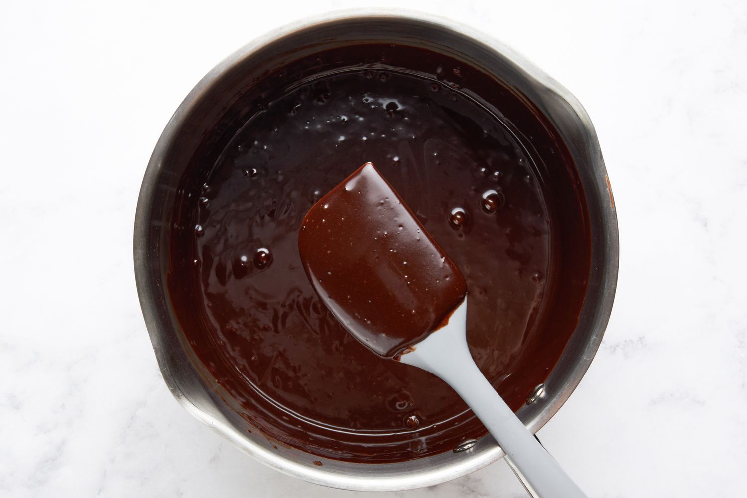 Hot fudge sauce on a spatula to showcase the desired texture and below the spatula, the saucepan with more hot fudge sauce