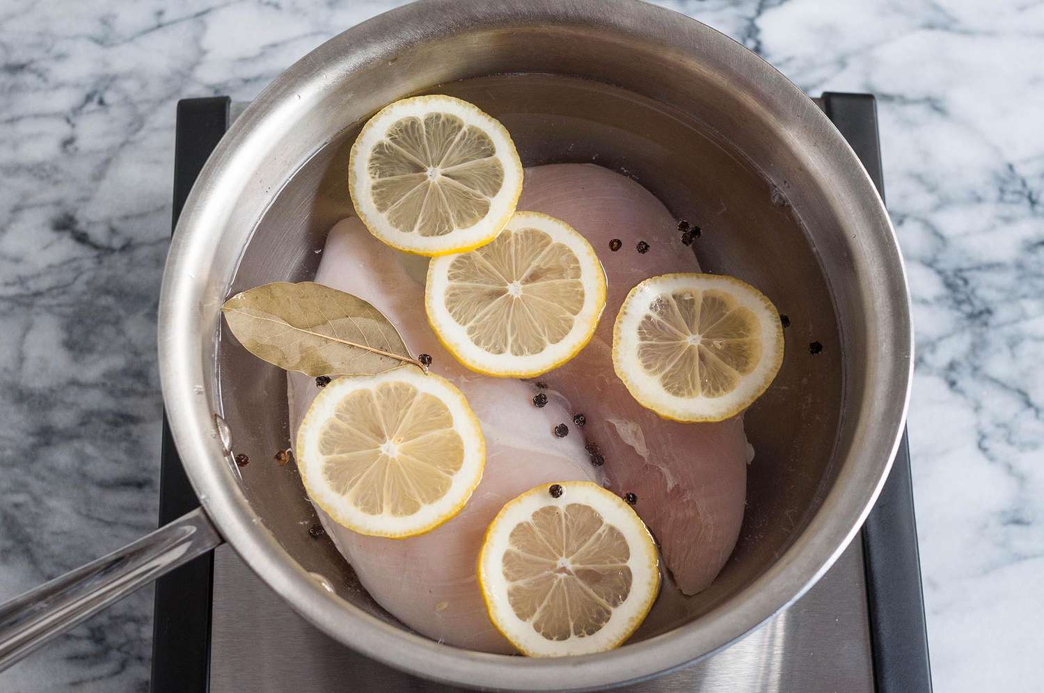 Two chicken breasts submerged in water in a pot with lemon slices on top
