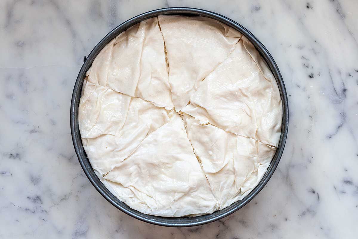 A cake pan is on a marble countertop. Unbaked phyllo dough is covering the top and cut into six pie shaped slices.