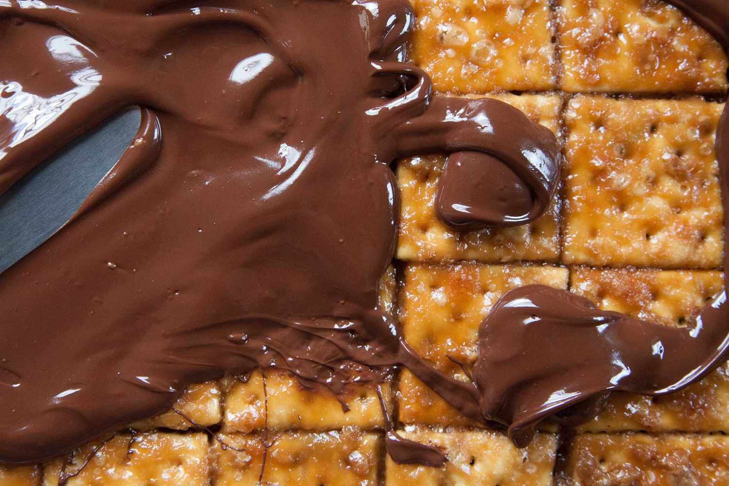 A chocolate mixture being spread over caramelized crackers with a butter knife