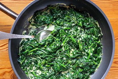 A skillet with Homemade Creamed Spinach and a serving spoon set on a wood table.