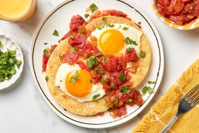 Huevos Rancheros Topped Chopped Cilantro on a Plate, and Surrounded by, a Small Plate of Chopped Cilantro, Orange Juice, a Bowl of Salsa, and a Deep Yellow Table Napkin With a Fork 